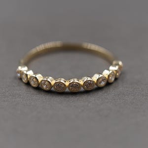 Puede incluir: Un anillo de oro que presenta una serie de pequeñas piedras redondas y transparentes engastadas en biseles individuales a lo largo de la mitad superior de la banda. Las piedras están dispuestas en una fila continua, creando un diseño circular.
