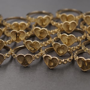 May include: A collection of gold-toned rings, each featuring a heart-shaped centerpiece with a textured surface and a single embossed capital letter. The bands have a delicate, linked chain design, and the rings are arranged in a group.