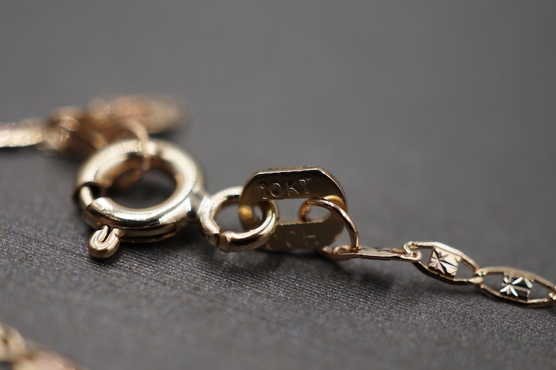 May include: Close-up of a gold chain necklace clasp with a 10KT marking. The chain is a delicate, patterned design.