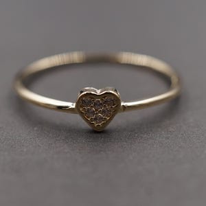 Puede incluir: Un delicado anillo de oro con un centro en forma de corazón. El corazón está adornado con pequeñas piedras brillantes. El anillo está sobre un fondo gris, resaltando su elegante diseño. Una joya.
