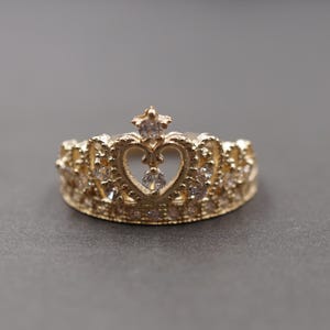 Könnte beinhalten: Ein goldfarbener Kronenring mit herzförmigem Mittelteil und kleinen, klaren Edelsteinen. Der Ring hat ein detailliertes Design mit einem sternförmigen Akzent oben. Der Ring ist vor einem grauen Hintergrund platziert.