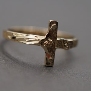 May include: A gold-toned ring with a detailed cross design. The ring band is textured with a diamond pattern.