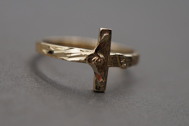 May include: A gold-toned ring with a detailed cross design. The ring band is textured with a diamond pattern.