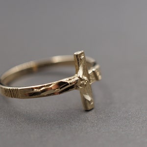 May include: A gold-toned ring with a detailed cross design. The ring band is textured with a diamond-cut pattern.