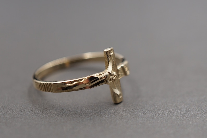 May include: A gold-toned ring with a detailed cross design. The ring band is textured with a diamond-cut pattern.