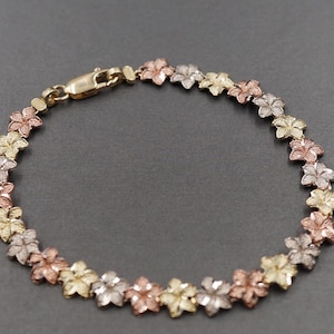 Könnte beinhalten: Ein zartes Armband mit floralem Design. Das Armband besteht aus kleinen, dreifarbigen Blumenanhängern in Gold, Roségold und Silber. Der Verschluss ist goldfarben. Der Schmuck wird auf einer grauen Oberfläche präsentiert.