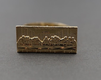 May include: A gold-colored ring featuring a rectangular plaque with a detailed relief of the Last Supper. The ring has a polished finish and is set against a dark gray background.