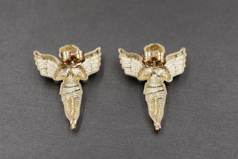 May include: A pair of gold angel wing earrings with a detailed design. The earrings are made of metal and have a post back closure.