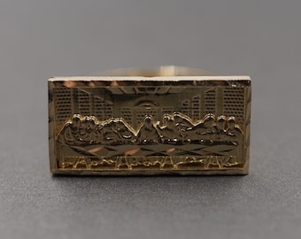 May include: A gold-colored ring with a detailed depiction of the Last Supper. The ring has a diamond-cut design on the band.