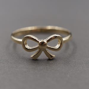 May include: A gold-coloured ring with a bow design. The bow is the central feature, with an intricate, openwork structure. The ring band is slender and plain, enhancing the bow's delicate form. The ring is set against a grey background.