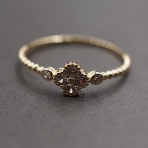 May include: A gold ring featuring a central four-petaled flower motif set with small clear stones. The band has a textured, beaded appearance, and two additional small clear stones are set on either side of the central floral design.