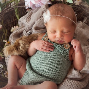 May include: A newborn baby in a sage green knitted romper with wooden buttons, resting in a nest of twigs, flowers, and a soft blanket. The baby wears a white floral headband. The scene is soft and natural.