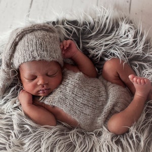 May include: A newborn baby wearing a gray knitted hat and matching outfit sleeps peacefully in a fluffy white and gray fur nest.