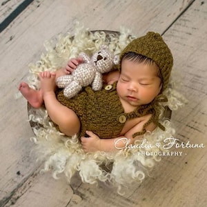 May include: A newborn baby wearing a brown knitted hat and matching outfit is sleeping in a white fluffy basket. The baby is holding a small brown teddy bear. The image includes the text "Claudia Fortuna Photography."