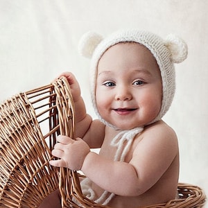 May include: A baby wearing a white knitted hat with bear ears sits in a wicker doll stroller with wooden wheels. The stroller is on a white wooden floor.