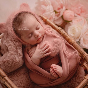 May include: A newborn baby girl sleeping in a wicker basket, wrapped in a pink swaddle and wearing a pink knitted bear hat. The basket is surrounded by pink and white flowers.