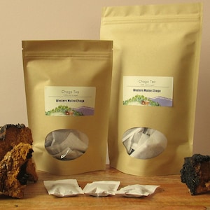 May include: Two brown paper bags with white labels that say "Chaga Tea 100% wild harvested" and "Western Maine Chaga". The bags contain tea bags and are sitting on a wooden surface with two pieces of chaga mushroom.