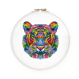 May include: A colorful cross-stitch pattern of a tiger's face, featuring vibrant hues of red, orange, yellow, green, blue, and purple. The design is set within a white embroidery hoop, creating a striking contrast. The artwork is a unique piece of textile art.