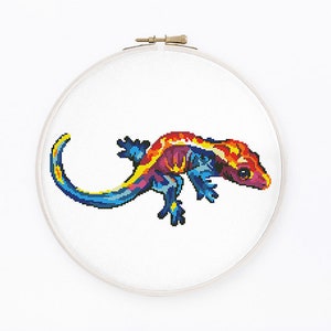 May include: A colourful cross-stitch pattern of a gecko with a blue tail, yellow and orange body, and pink head. The gecko is stitched on white fabric and framed in a wooden embroidery hoop.