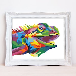 May include: A framed cross-stitch depicting a colourful iguana. The artwork uses a vibrant palette of blues, greens, yellows, and purples, in a pop art style. The iguana is set against a white background within a white ornate frame.