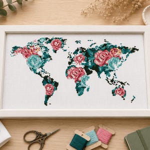 以下が含まれることがあります： 花柄のデザインが施された、額入りのクロスステッチの世界地図。 地図はピンクとティールブルーの花で埋め尽くされ、白い背景が特徴です。 フレームは白で、木製の表面に飾られています。