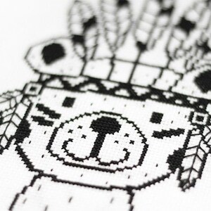 May include: Black and white cross-stitch pattern of a bear wearing a headdress with feathers. The bear has a simple, friendly face.