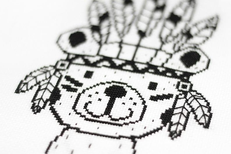 May include: Black and white cross-stitch pattern of a bear wearing a headdress with feathers. The bear has a simple, friendly face.