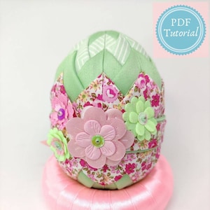 May include: A green and pink fabric Easter egg with a floral pattern and a pink satin base. The egg is decorated with pink and green fabric flowers. The image includes the text "Ada Quilted Creations" and "PDF Tutorial".