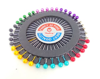 May include: A black pincushion with a circular design and a label that reads "Fancy 40 Pins Tulip Japan Best Quality". The pincushion is filled with colorful pins in a rainbow pattern.