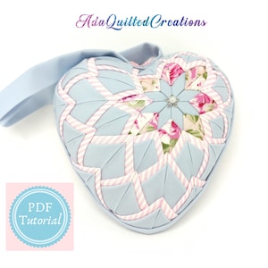 Op de afbeelding: Een lichtblauw, hartvormig, gestikt kussen met een roze-wit gestreept patroon en een bloemmotief in het midden. Het kussen heeft een lus om op te hangen en de tekst "Ada Quilted Creations" is zichtbaar op de achtergrond.