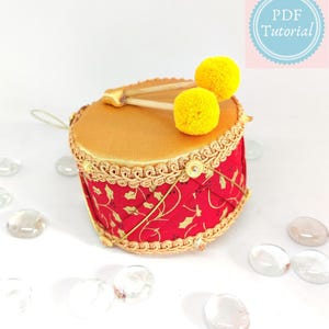 May include: A decorative drum ornament with a red and gold design. The drum features a red fabric with a gold floral pattern and gold trim. Two yellow pom-pom drumsticks rest on top. The image includes the text "PDF Tutorial".