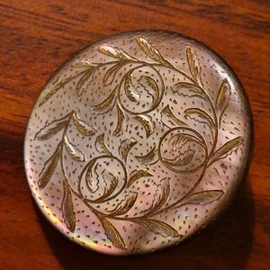 Puede incluir: Un estuche compacto redondo plateado con un diseño floral grabado. El estuche compacto tiene un brillo nacarado y está decorado con hojas y patrones florales arremolinados. El fondo es una superficie de madera oscura.