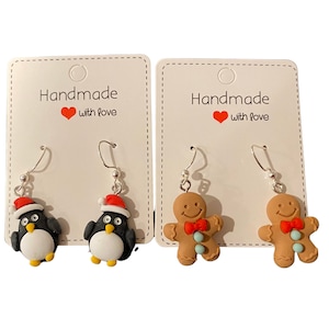 May include: Two pairs of handmade Christmas earrings. One pair features a penguin wearing a Santa hat and the other pair features a gingerbread man. Both pairs are on white cards with the text "Handmade with love".