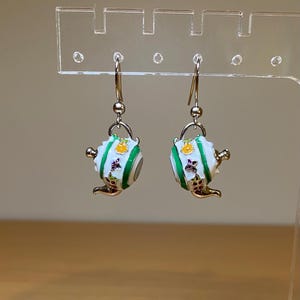 May include: A pair of silver dangle earrings with white teapot charms. The teapots have green stripes, yellow accents, and purple and white floral details.