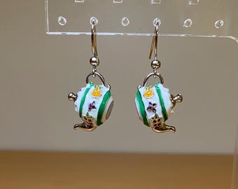 May include: A pair of silver dangle earrings with white teapot charms. The teapots have green stripes, yellow accents, and purple and white floral details.