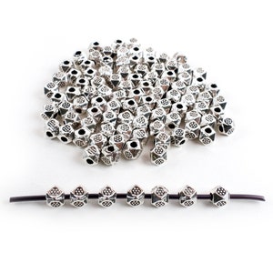 May include: A large pile of silver metal beads with a geometric design. The beads are arranged in a loose pile on a white background. A smaller row of the same beads is strung on a black cord.