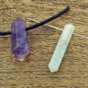 May include: Two crystal pendants, one purple amethyst and one green aventurine, are strung on a black cord. The crystals are both pointed at the bottom and have a smooth, polished finish.