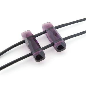 May include: Two purple crystal beads on a black leather cord. The beads are rectangular and have a faceted design.