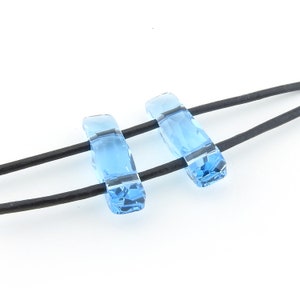 May include: Two light blue crystal beads on a black leather cord. The beads are rectangular and have a faceted design.