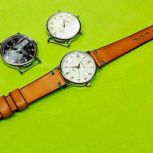 May include: A brown leather watch strap with blue stitching and three silver watches. Two of the watches have white faces and the third has a black face.