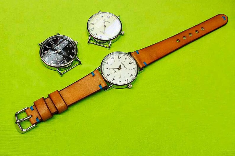 May include: A brown leather watch strap with blue stitching and three silver watches. Two of the watches have white faces and the third has a black face.