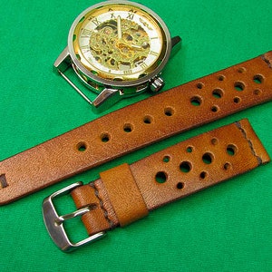May include: A brown leather watch strap with a perforated design. The strap has a silver buckle and is shown on a green surface.