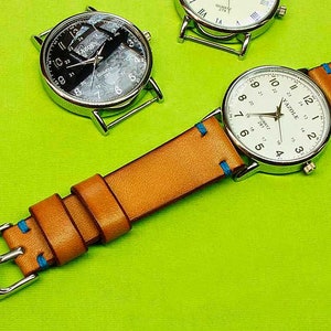 May include: A silver watch with a white face and black numbers. The watch has a brown leather band with blue stitching. The watch is on a green surface.