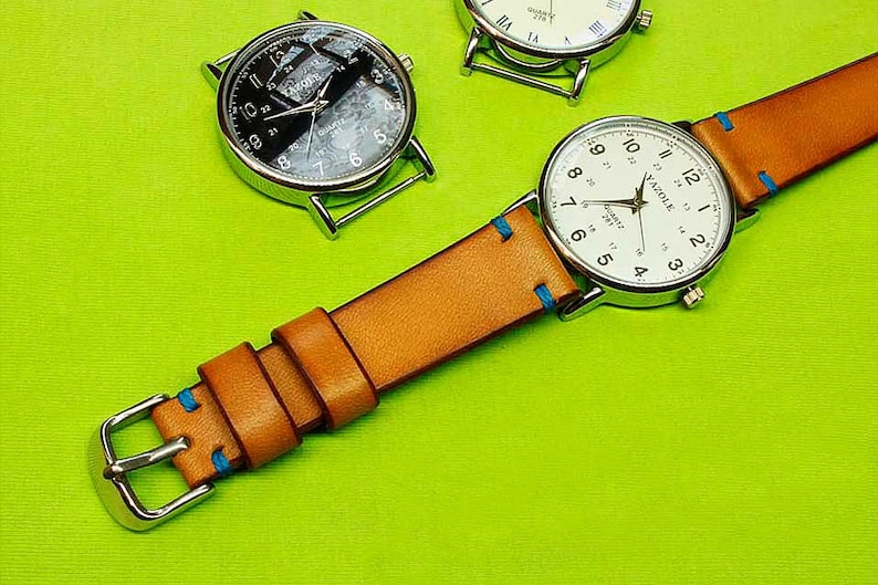 May include: A silver watch with a white face and black numbers. The watch has a brown leather band with blue stitching. The watch is on a green surface.