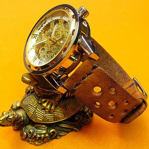 May include: A gold-toned wristwatch with a skeletonized dial and a brown leather strap. The watch is displayed on a gold-colored turtle figurine.