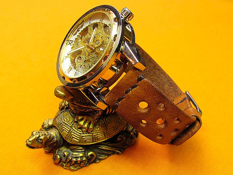 May include: A gold-toned wristwatch with a skeletonized dial and a brown leather strap. The watch is displayed on a gold-colored turtle figurine.