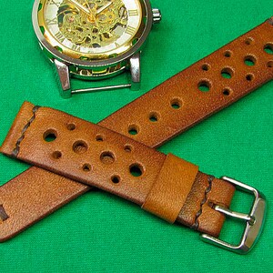 May include: A brown leather watch strap with a perforated design and a silver buckle. The strap is shown on a green surface.