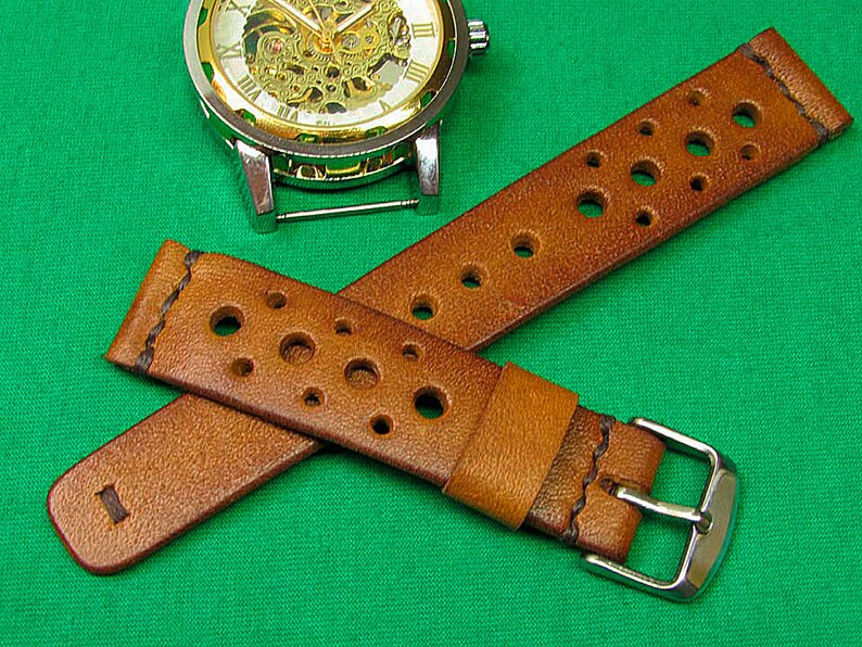 May include: A brown leather watch strap with a perforated design and a silver buckle. The strap is shown on a green surface.