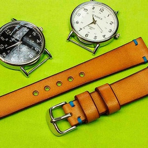 May include: Two leather watch straps with blue stitching. The straps are tan in color and have a buckle on one end. The straps are laid out on a green surface.