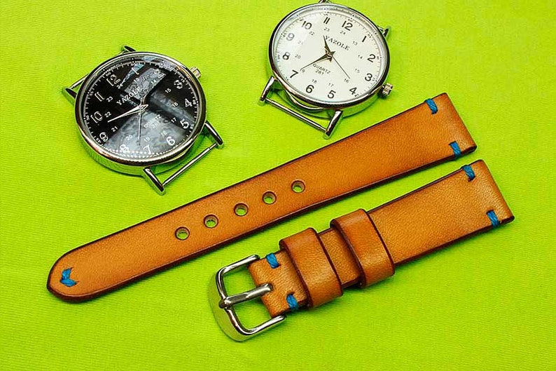 May include: Two leather watch straps with blue stitching. The straps are tan in color and have a buckle on one end. The straps are laid out on a green surface.